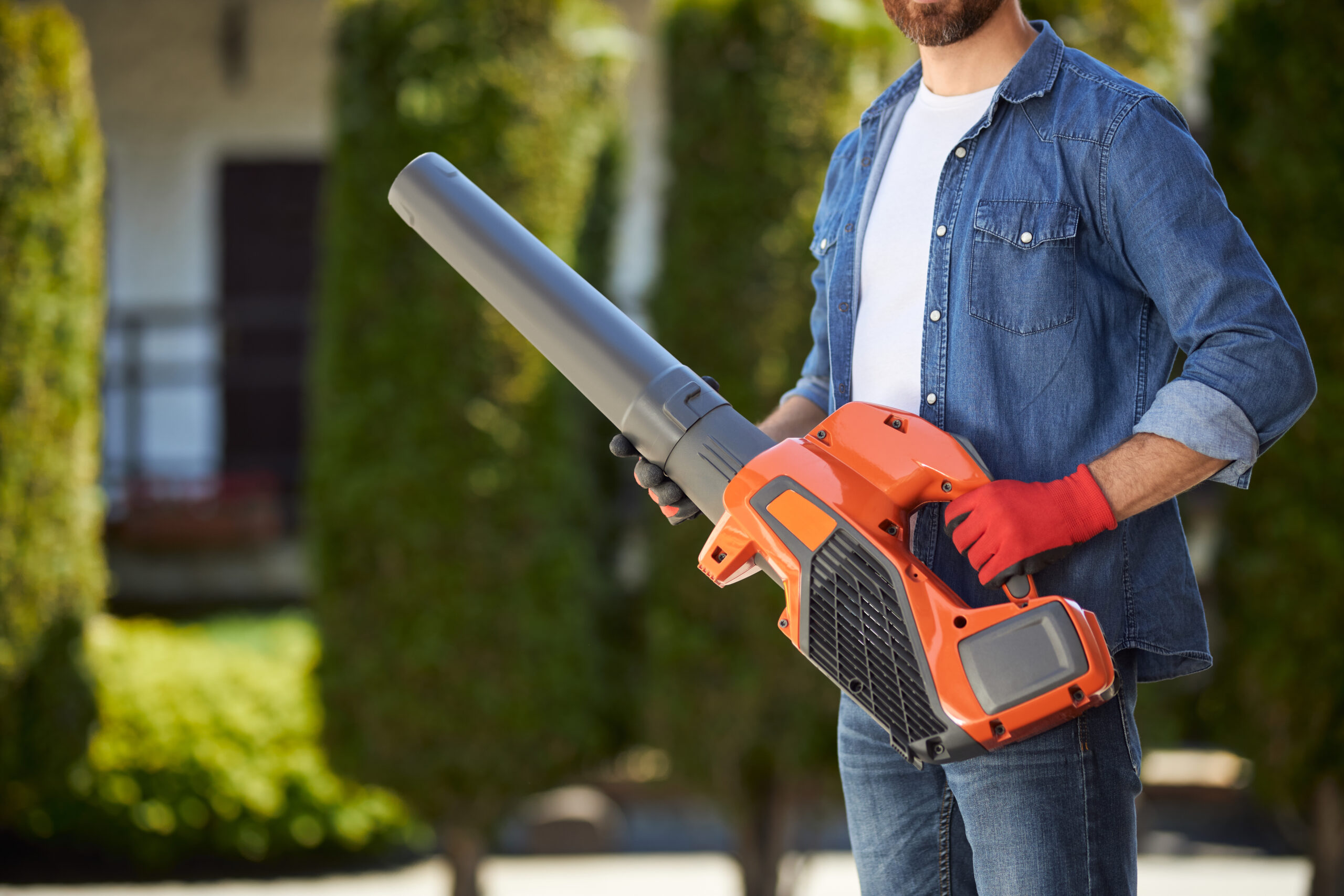 Bearded male gardener in gloves holding powerful cordless leaf blower outdoors. Crop view of handyman in denim shirt using modern garden equipment against blurred background. Concept of seasonal work.